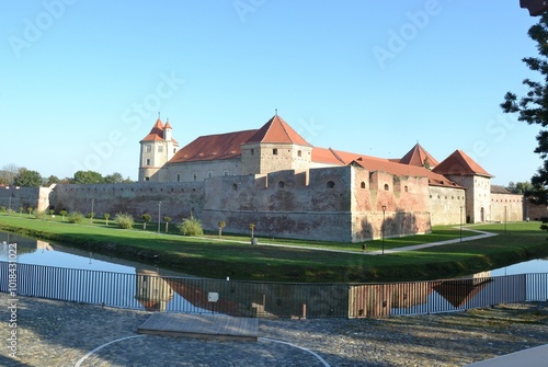 This photo depicts the impressive Făgăraș Fortress in Romania. The medieval structure, with its thick stone walls and red-tiled roofs, is reflected in the surrounding moat,