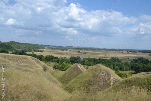 
The photo captures the rolling hills and open plains of Mogrytsia in the Sumy region of Ukraine. The serene landscape features grassy slopes, scattered trees, and a vast blue sky.