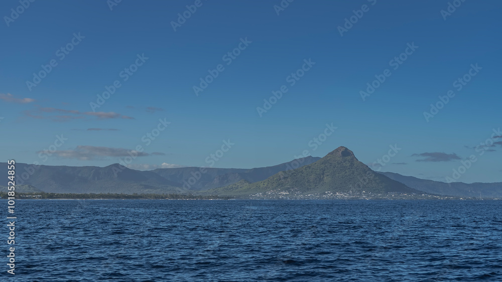 The landscape of the coast of a tropical island. Picturesque mountains covered with green vegetation, against a blue sky and clouds. City buildings on the shore. In the foreground is the ocean. The landscape of the coast of a tropical island. Picturesque mountains covered with green vegetation, against a blue sky and clouds. City buildings on the shore. In the foreground is the ocean.