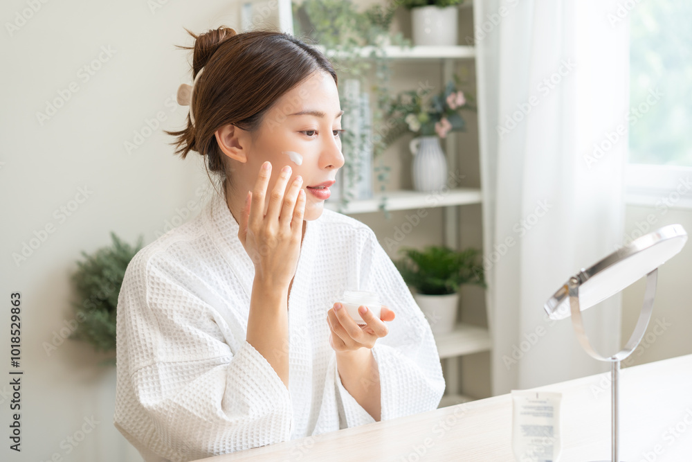 Facial beauty skin care, smile of pretty asian young woman in bathrobe looking at mirror, hand applying moisturizer lotion on her face, putting cream treatment before makeup cosmetic routine at home. Facial beauty skin care, smile of pretty asian young woman in bathrobe looking at mirror, hand applying moisturizer lotion on her face, putting cream treatment before makeup cosmetic routine at home.