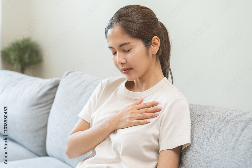 Heart attack, health care concept, suffering acute cardiac asian young woman hand touching on her chest pain, breathing problem, asthma attack, pressure while sitting on sofa with closed eyes at home. Heart attack, health care concept, suffering acute cardiac asian young woman hand touching on her chest pain, breathing problem, asthma attack, pressure while sitting on sofa with closed eyes at home.