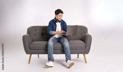 Full body image of young Asian man using laptop sitting on sofa on white background