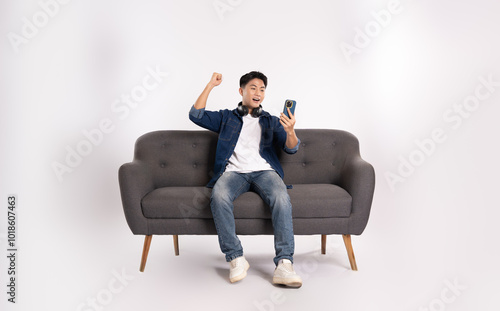 Full body image of young Asian man using phone sitting on sofa on white background