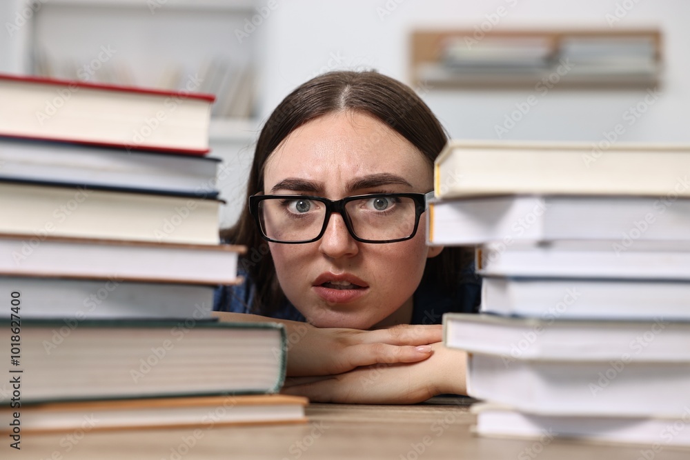 Worried student before exam at table among stacks of books indoors Worried student before exam at table among stacks of books indoors