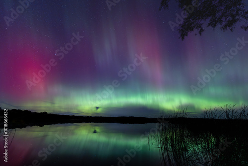 Northern Lights erupt over a Minnesota lake in the dark sky overhead shining a bright rainbow of Aurora light and colors over the forests and clean water during strong solar storm