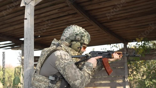 A soldier in full combat gear aims his assault rifle while taking cover behind a wooden structure. The tactical environment suggests an intense military operation or training exercise in a close-quart