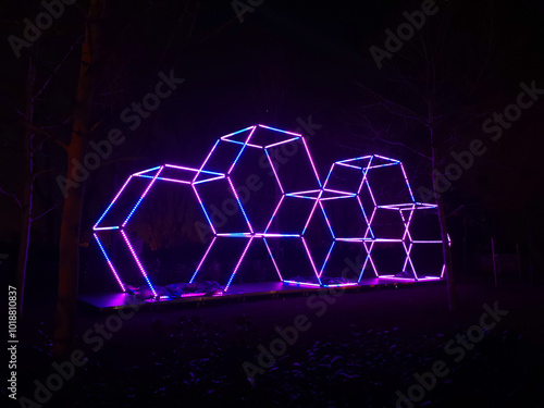 A futuristic geometric structure glows with vibrant purple and blue lights, illuminating the dark outdoor environment. photo