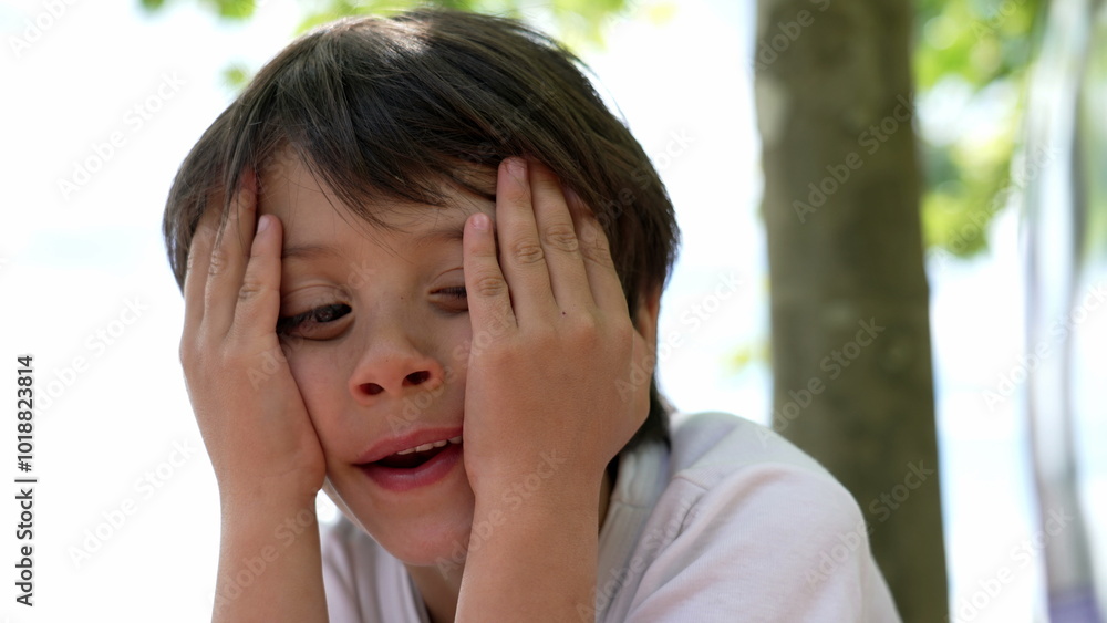 Young boy resting his head in his hands, looking tired but content, seated outdoors on a sunny day, with a soft background of trees and natural light Young boy resting his head in his hands, looking tired but content, seated outdoors on a sunny day, with a soft background of trees and natural light