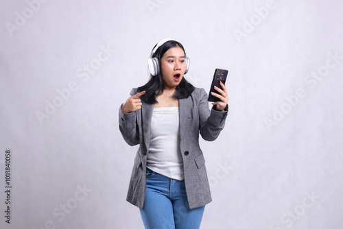 beautiful adult office lady shocked to cellphone wearing headphone and holding mobile phone while pointing wearing suit isolated on white background. lifestyle, education, entertainment concept