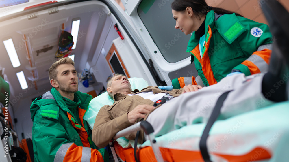 Paramedics in green and orange uniforms attend to a male patient on a stretcher, preparing him for transport inside an ambulance. The scene captures emergency medical care and teamwork in action. Paramedics in green and orange uniforms attend to a male patient on a stretcher, preparing him for transport inside an ambulance. The scene captures emergency medical care and teamwork in action.