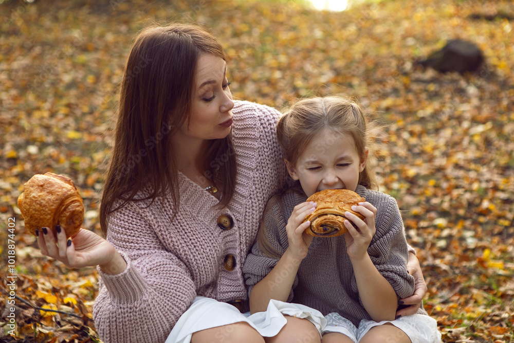mother and her daughter of seven years walk warm autumn in a park with trees at sunset in orange leaves. sitting on the ground eating croissants.