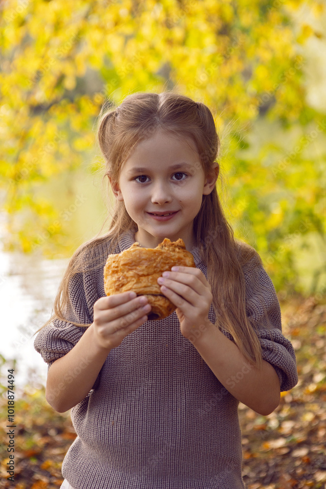 A girl of seven holds a curroissant bun in the park at sunset in autumn