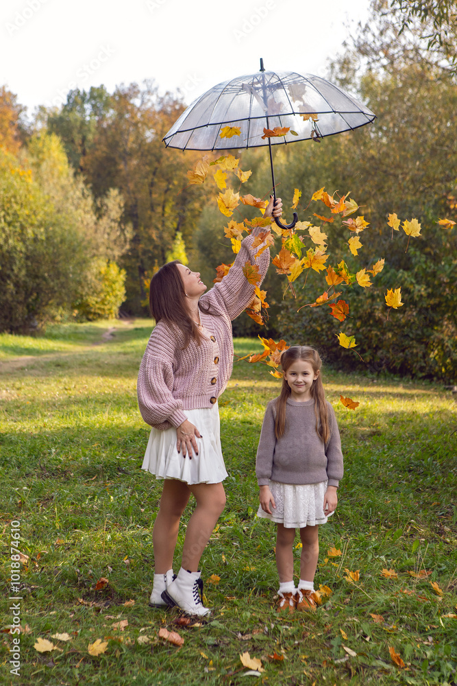 mother and her daughter of seven years walk warm autumn in a park with trees at sunset in orange leaves. holding a transparent umbrella with leaves falling out of it