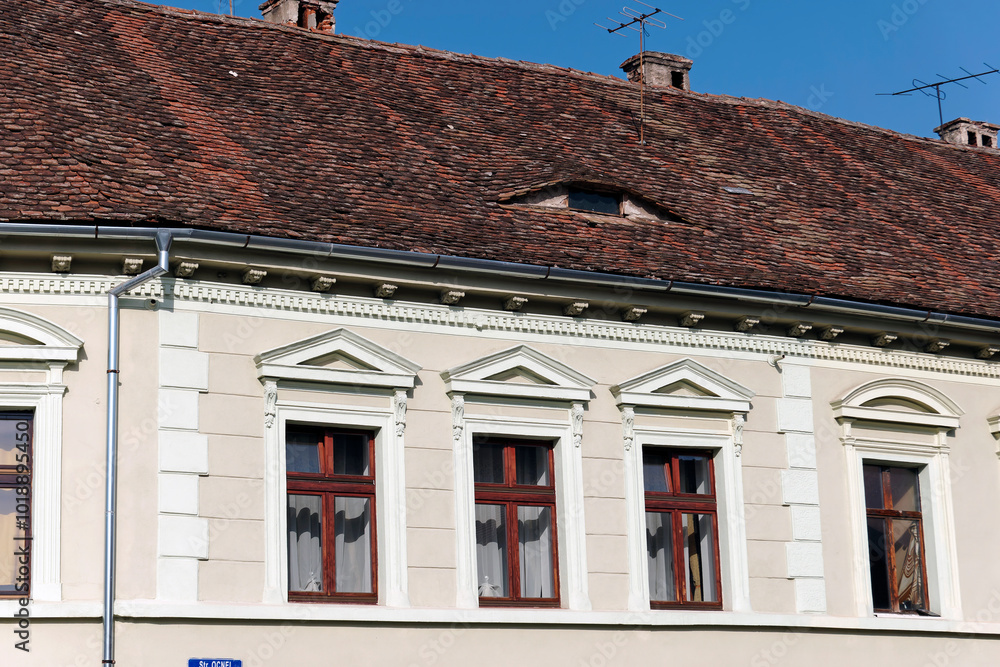 A close-up view of a traditional building facade in Sibiu, Romania, featuring a red tiled roof with a distinctive arched shape. The building also has several window with white trim and brown frames. A close-up view of a traditional building facade in Sibiu, Romania, featuring a red tiled roof with a distinctive arched shape. The building also has several window with white trim and brown frames.