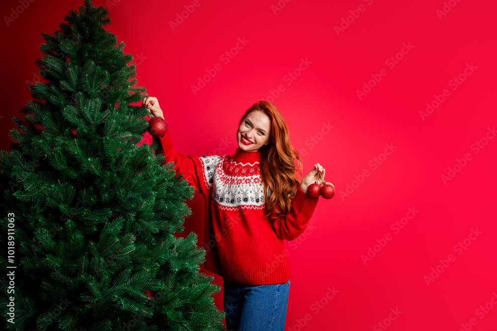 Photo portrait of cheerful positive young girl wearing ugly sweater decorating xmas fir tree with toys balls isolated on red color background Photo portrait of cheerful positive young girl wearing ugly sweater decorating xmas fir tree with toys balls isolated on red color background