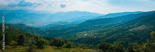 A breathtaking view of verdant valleys and distant mountains bathed in sunlight, under a sky dotted with clouds.