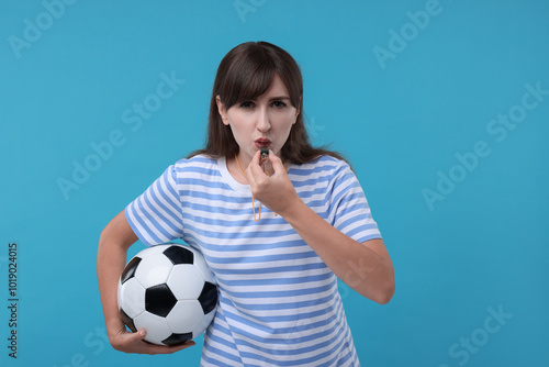 Woman with soccer ball blowing whistle on light blue background