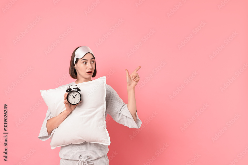 Overslept young woman with sleep mask, pillow and alarm clock pointing at something on pink background. Space for text Overslept young woman with sleep mask, pillow and alarm clock pointing at something on pink background. Space for text
