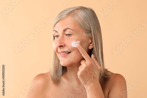 Senior woman applying face cream on beige background