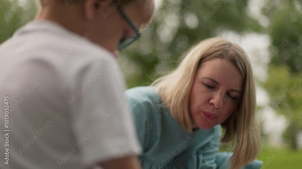 A woman wearing a blue dress is seen blowing air at something intently while a blurry view of a child wearing glasses appears in the background A woman wearing a blue dress is seen blowing air at something intently while a blurry view of a child wearing glasses appears in the background