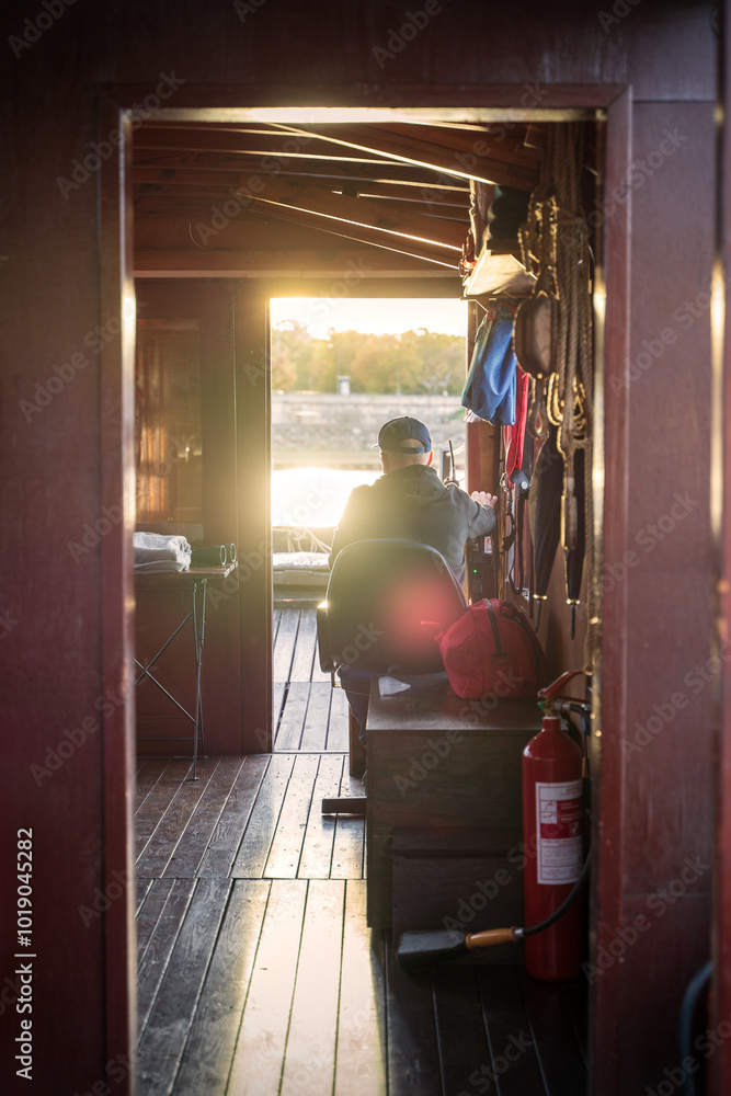 A man is steering a boat during sunset, viewed from inside the cabin. The warm glow of the sun reflects off the wooden interior, creating a serene, peaceful atmosphere on the water.
