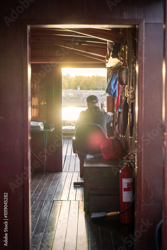 Obraz na płótnie A man is steering a boat during sunset, viewed from inside the cabin. The warm glow of the sun reflects off the wooden interior, creating a serene, peaceful atmosphere on the water. w salonie