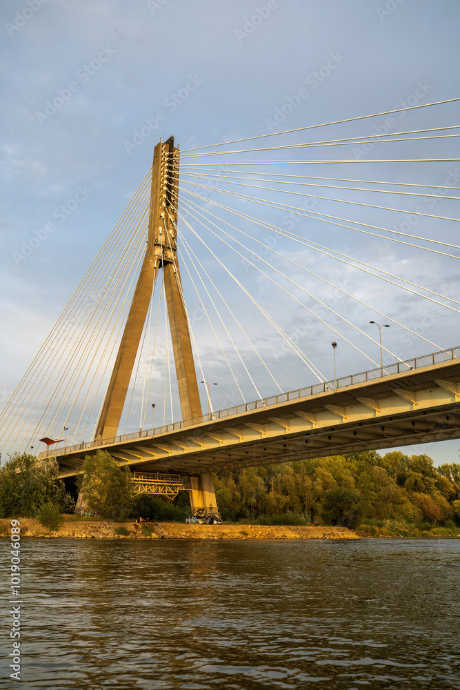 A suspension bridge with cables stretching across a river under a clear sky. The bridge stands tall, reflecting golden light, surrounded by trees and water below. Warsaw, Poland