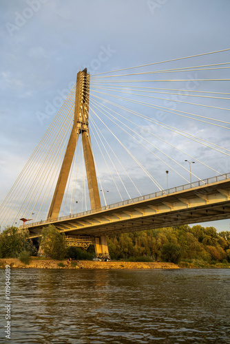 Obraz na płótnie A suspension bridge with cables stretching across a river under a clear sky. The bridge stands tall, reflecting golden light, surrounded by trees and water below. Warsaw, Poland w salonie