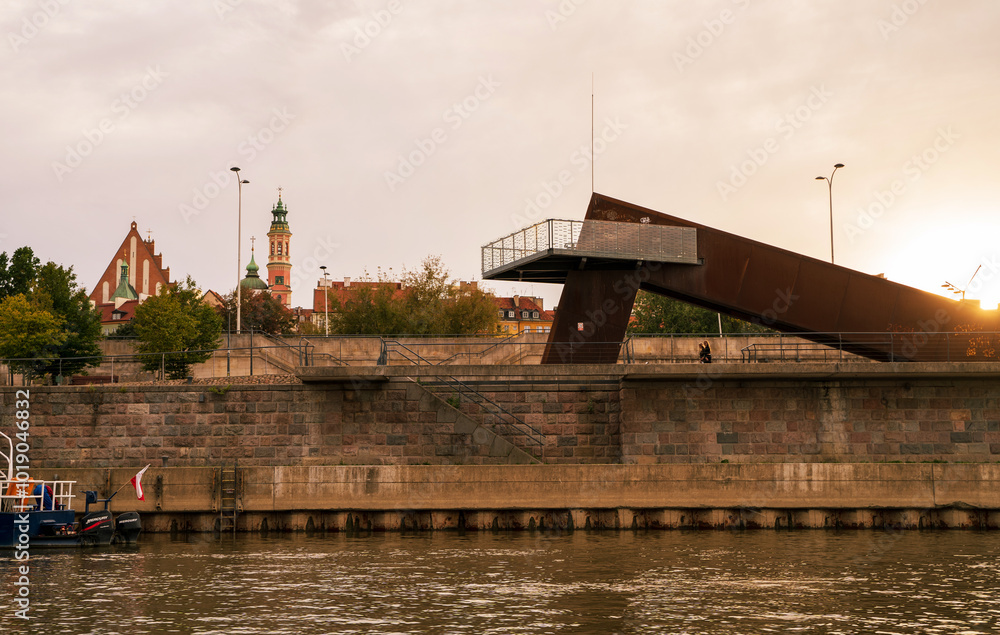 A modern observation deck with a rust-colored metal structure juts out over the Vistula River in Warsaw, Poland. The city skyline, featuring historic churches and buildings