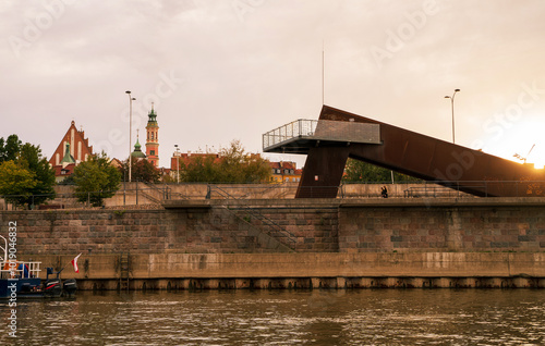Obraz na płótnie A modern observation deck with a rust-colored metal structure juts out over the Vistula River in Warsaw, Poland. The city skyline, featuring historic churches and buildings w salonie