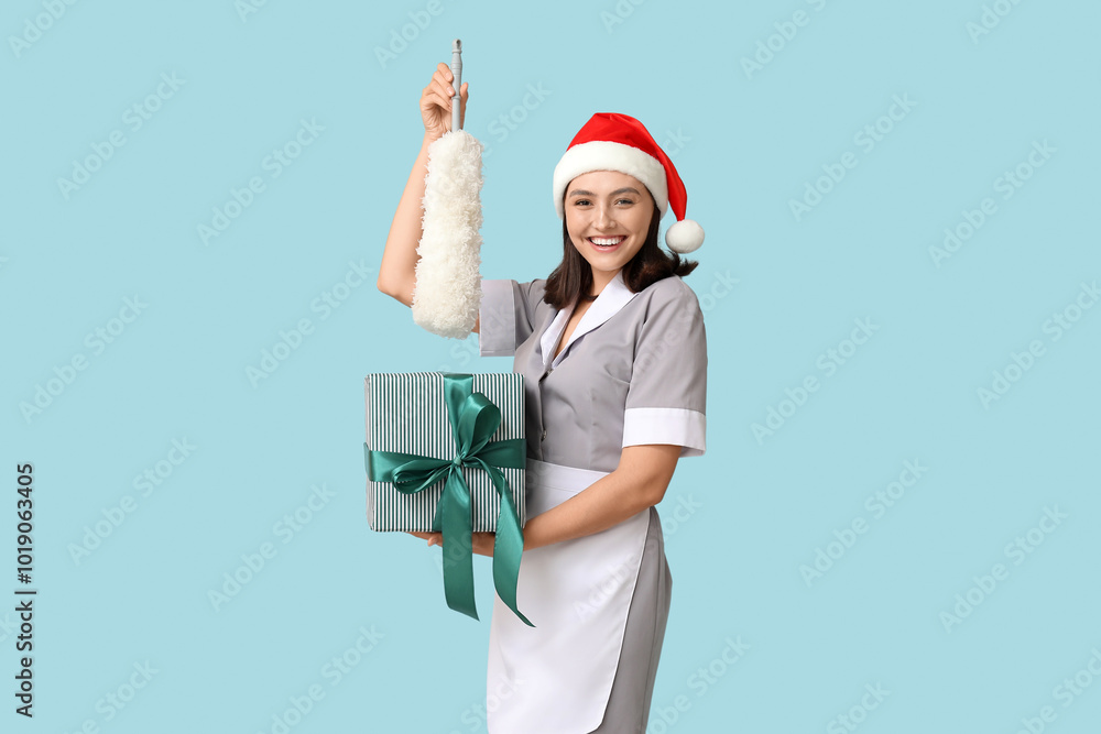 Beautiful female chambermaid in Santa hat with pp-duster and gift box on blue background Beautiful female chambermaid in Santa hat with pp-duster and gift box on blue background