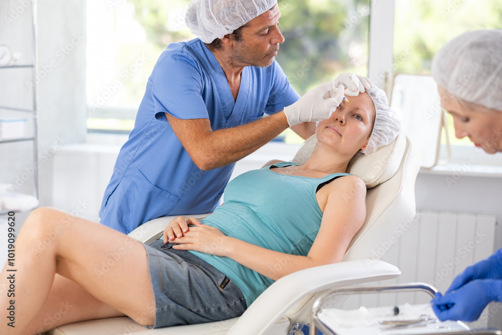 Obraz premium Male doctor with help of mature nurse assistant performs rejuvenation procedure for female client and gives injection of botulinum toxin in forehead and bridge of nose