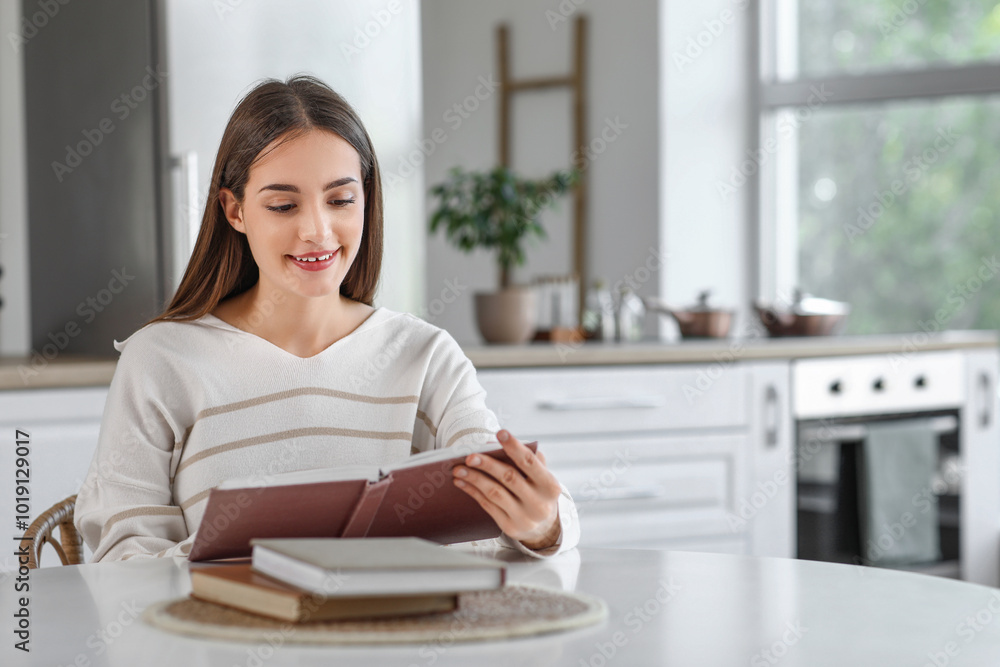 Beautiful young happy woman reading book at table in kitchen Beautiful young happy woman reading book at table in kitchen