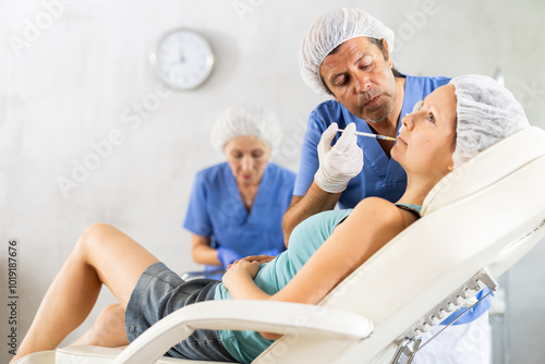 Man performs rejuvenating injection to smooth out wrinkles in area around mouth to female patient, mature female nurse assists, conducts preparatory manipulations photo