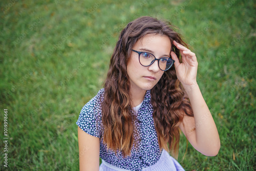 Portrait of girl in glasses with curly hair sitting in park on grass. Concept of learning in nature Portrait of girl in glasses with curly hair sitting in park on grass. Concept of learning in nature