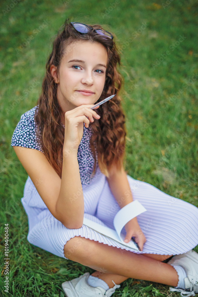 Portrait of cute smiling girl in glasses with curly hair sitting in park on grass. Concept of learning in nature Portrait of cute smiling girl in glasses with curly hair sitting in park on grass. Concept of learning in nature