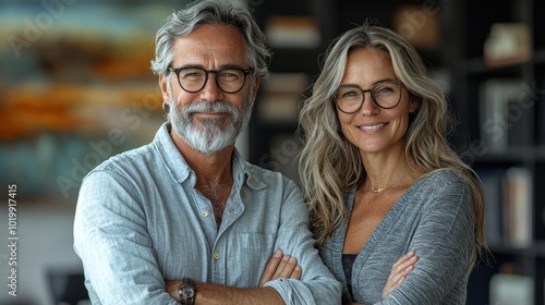 A gray-haired man and a woman with long hair, both in glasses, smiling warmly with arms crossed in a well-lit interior space, suggesting professionalism and warmth. photo