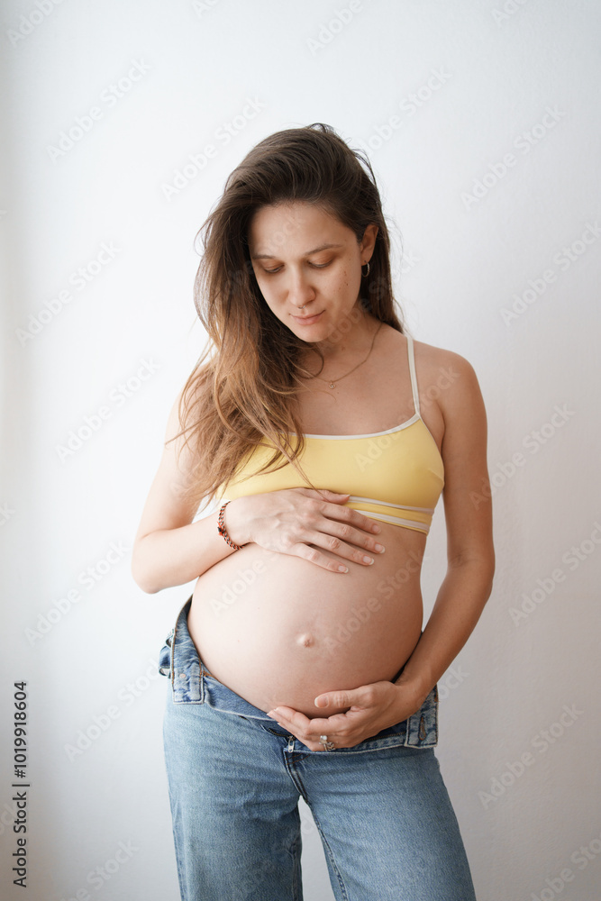 Pregnant woman in yellow top gently touching her belly near a window Pregnant woman in yellow top gently touching her belly near a window