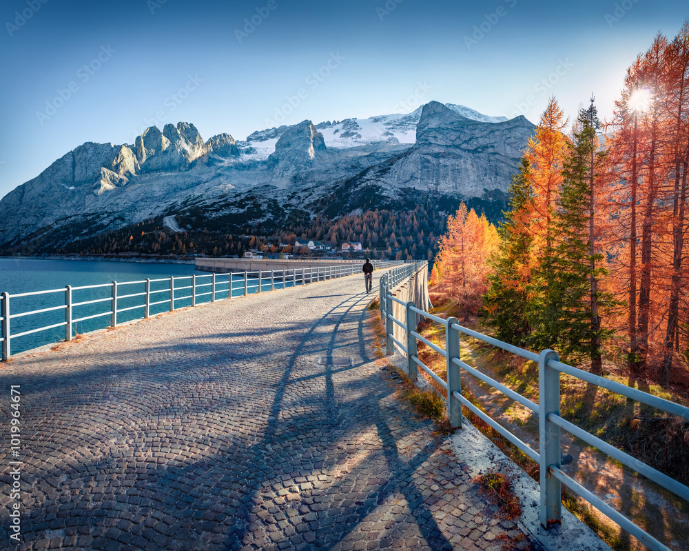 Tourist walks on dam on Fedaia lake, Italy, Europe. Stunning autumn view of Dolomites. Fantastic morning scene of Italian Alps. Traveling concept background.