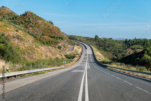 Estrada com uma curva ao fundo, com as barras metálicas de proteção e duas linhas continuas ao centro em meio a uma zona montanhosa e um céu azul photo