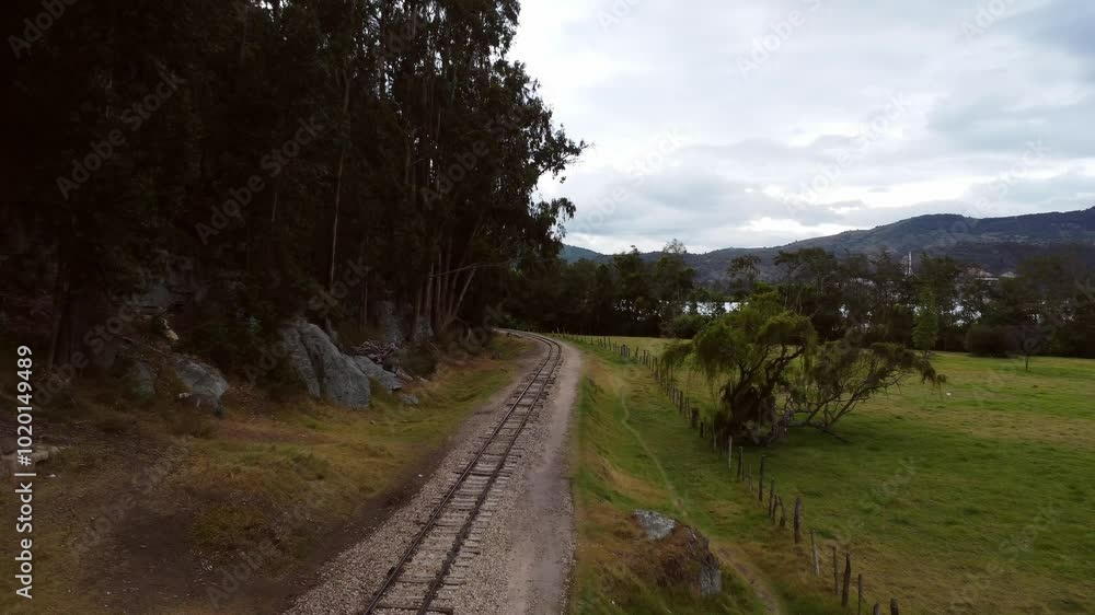 A deserted railway track runs through a quiet green field with mountains in the background