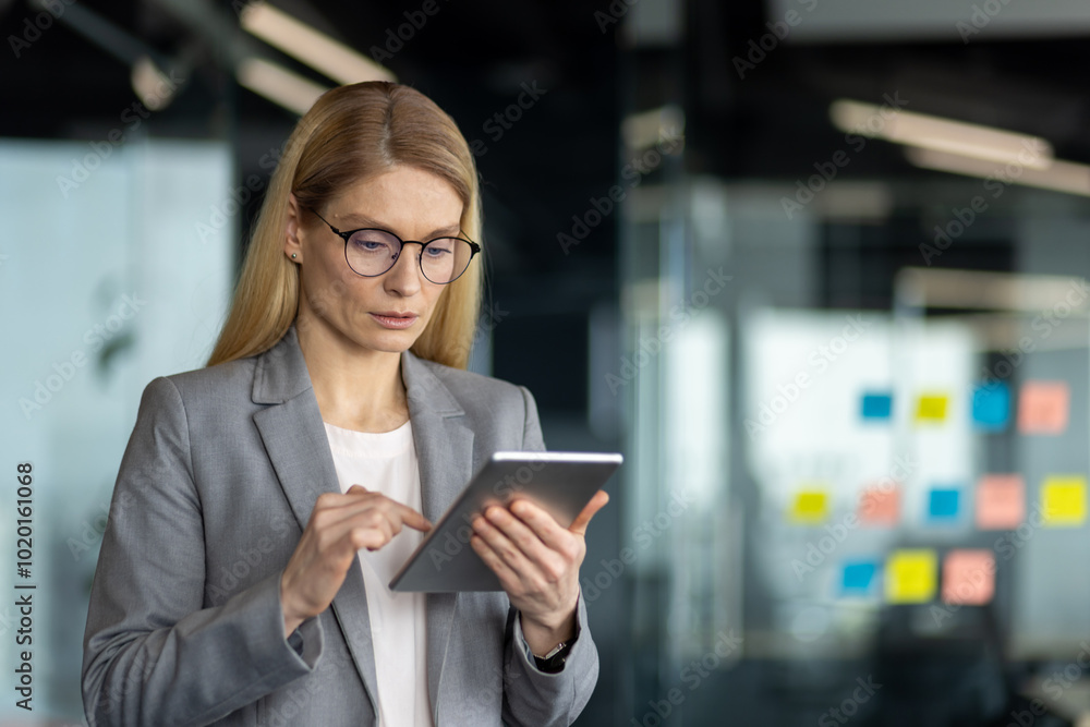 Confident businesswoman in gray suit using digital tablet in bright modern office. She looks focused, embodying professionalism and technology-driven work environment. Confident businesswoman in gray suit using digital tablet in bright modern office. She looks focused, embodying professionalism and technology-driven work environment.