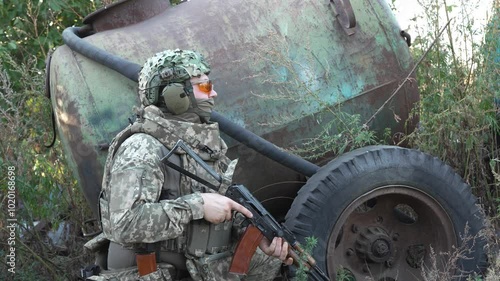 A soldier in camouflage aims a rifle while taking cover behind rusty equipment, focused on a mission in a rural combat zone. The scene captures the tension and dedication of military operations