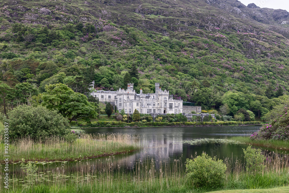 A scenic view of Kylemore Abbey in Connemara, Ireland, nestled among dense forests and rugged hills by Pollacapall Lough A scenic view of Kylemore Abbey in Connemara, Ireland, nestled among dense forests and rugged hills by Pollacapall Lough