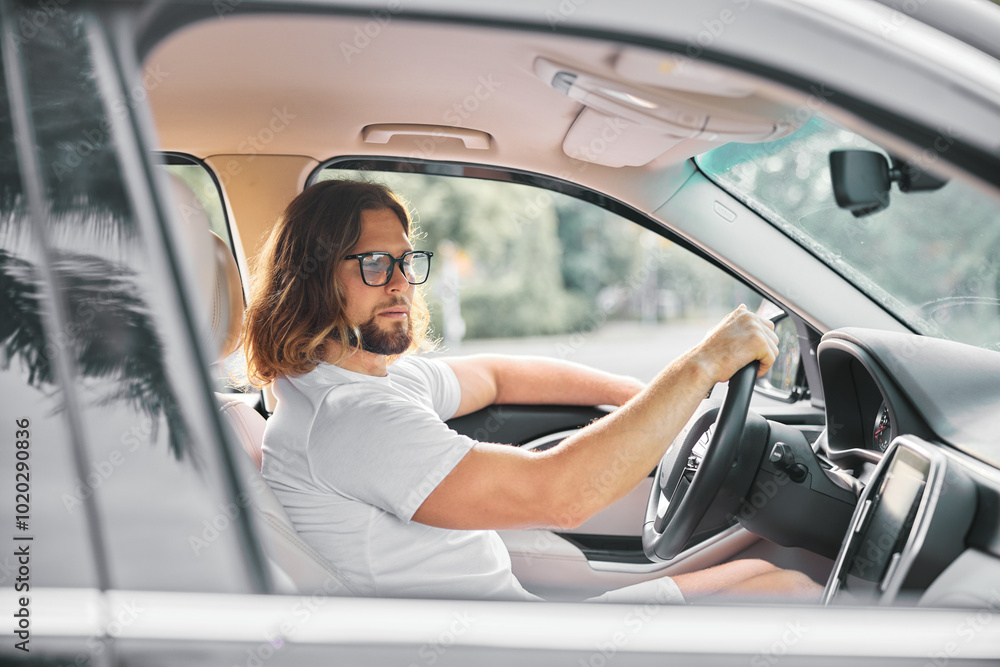 Man driving a car with a relaxed expression and sunglasses, seated on the driver s side, trees visible in the background Man driving a car with a relaxed expression and sunglasses, seated on the driver s side, trees visible in the background