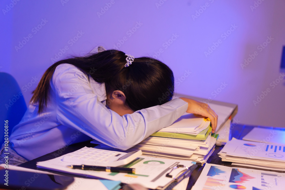 A young woman works overtime at night at her desk in office, using a laptop reviewing documents. is stressed, tired,experiencing headaches,body aches from sitting too long,leading to office syndrome A young woman works overtime at night at her desk in office, using a laptop reviewing documents. is stressed, tired,experiencing headaches,body aches from sitting too long,leading to office syndrome
