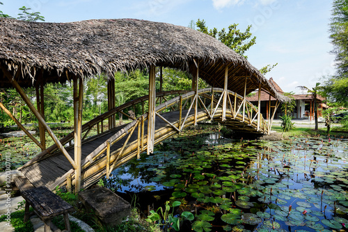 A curved bamboo bridge with a thatched leaf roof crosses a small lake covered with lotus plants photo