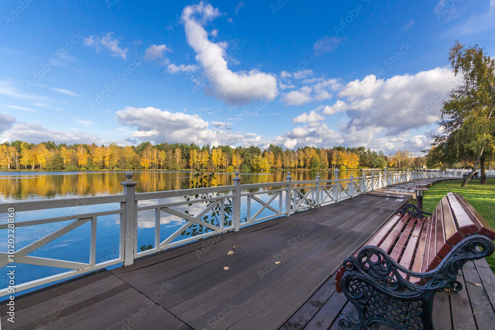 Obraz premium Wooden river embankment with white fences. Metal frame of a bench with a wooden seat. Beautiful landscape on a sunny day.
