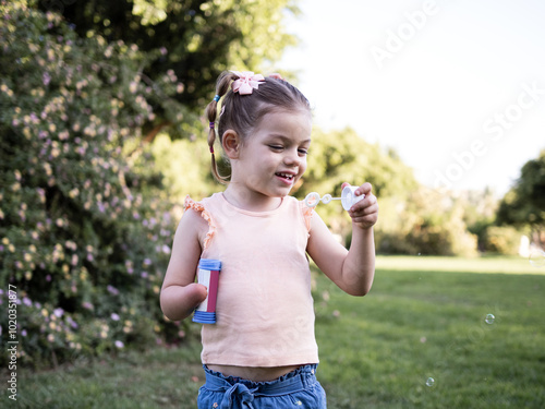 A happy young girl with disability blows bubbles in a green park, holding a bubble wand and a container. photo