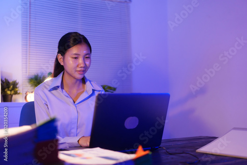 An Asian woman is working overtime at her desk in the office, busy viewing documents and her notebook. She is talking while managing tasks late at night, staying focused and productive